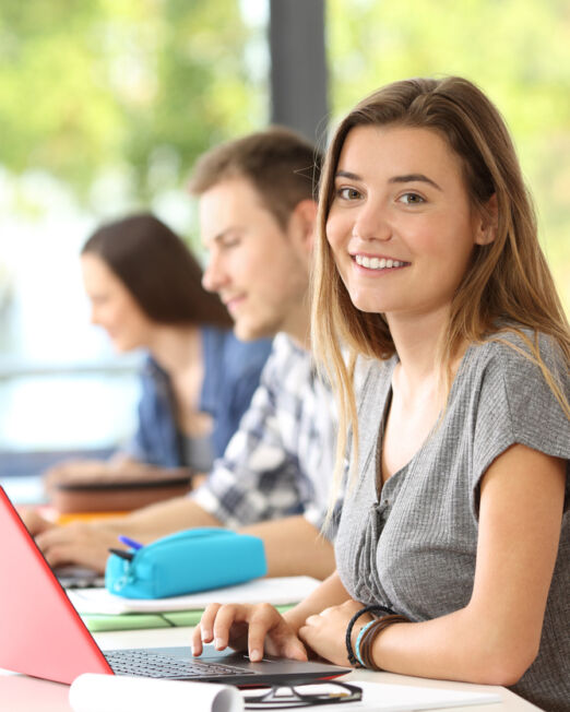 Happy student posing in a classroom looking at you with an unfocused window in the background